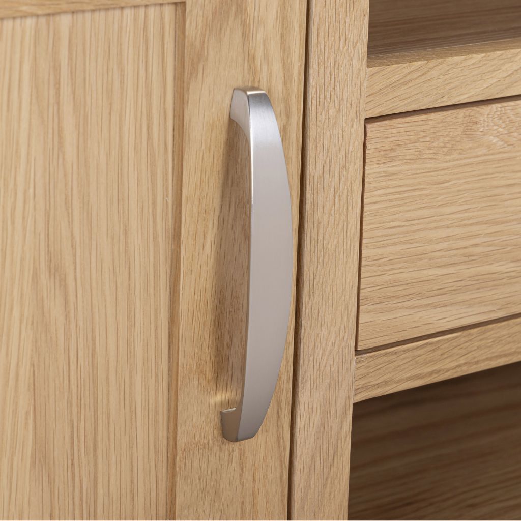 A curved, brushed silver metal cabinet handle mounted on a light oak wood door next to a matching drawer.